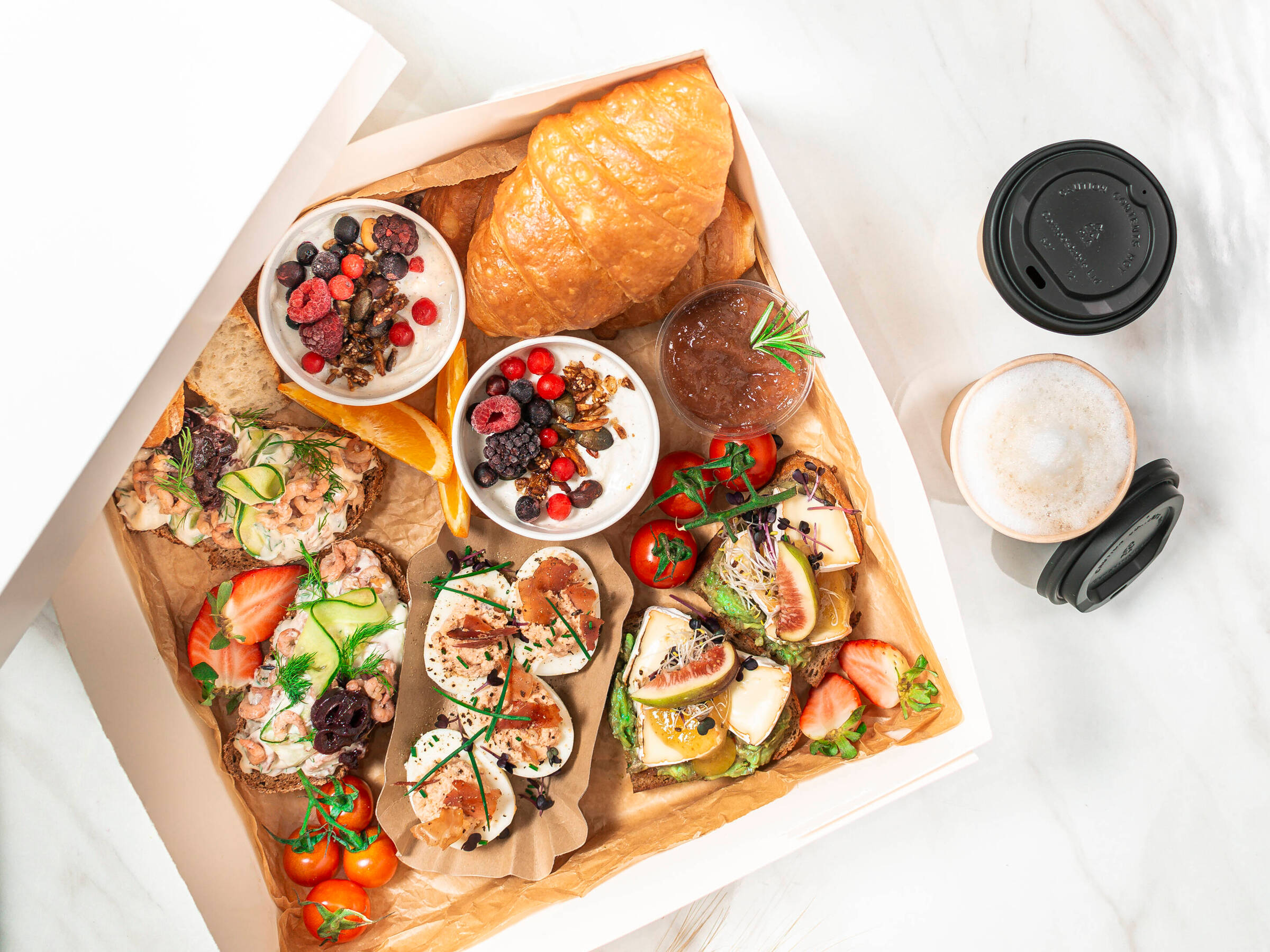 Eine weiße Frühstücksbox mit zweimal Porridge mit beeren, Croissant, Marmelade und einem Coffee to go.